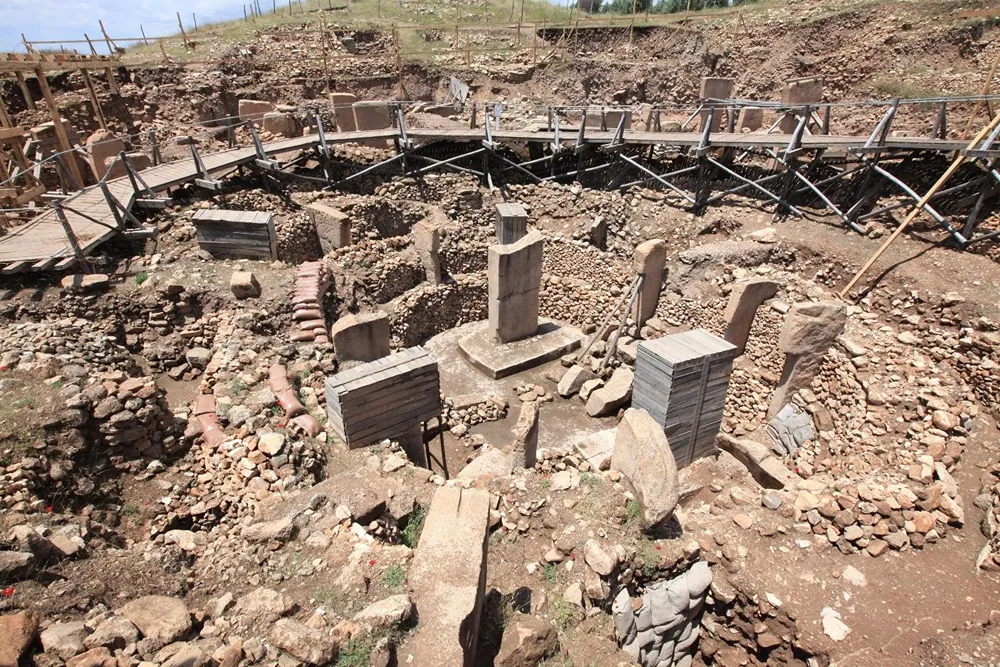 Arkeolojik kazı alanında ortaya çıkarılmış taş yapılar ve dikili taşlar; çevresinde ahşap yürüyüş yolları bulunuyor