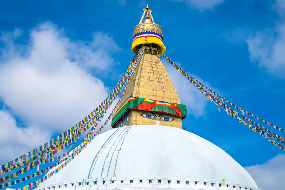Mavi gökyüzü altında, Buda’nın gözlerini taşıyan beyaz kubbeli bir stupa ve renkli dua bayrakları