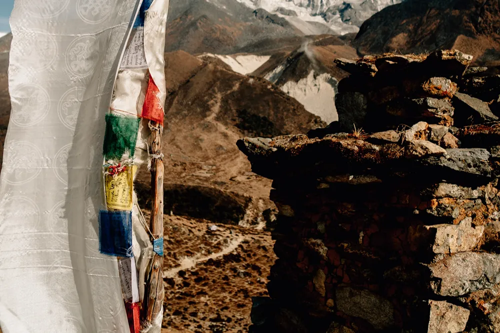 nepal-resimler (31) Dağlık vadide, taş bir yapının yanında renkli Tibet dua bayrakları ve karla kaplı zirveler görülüyor