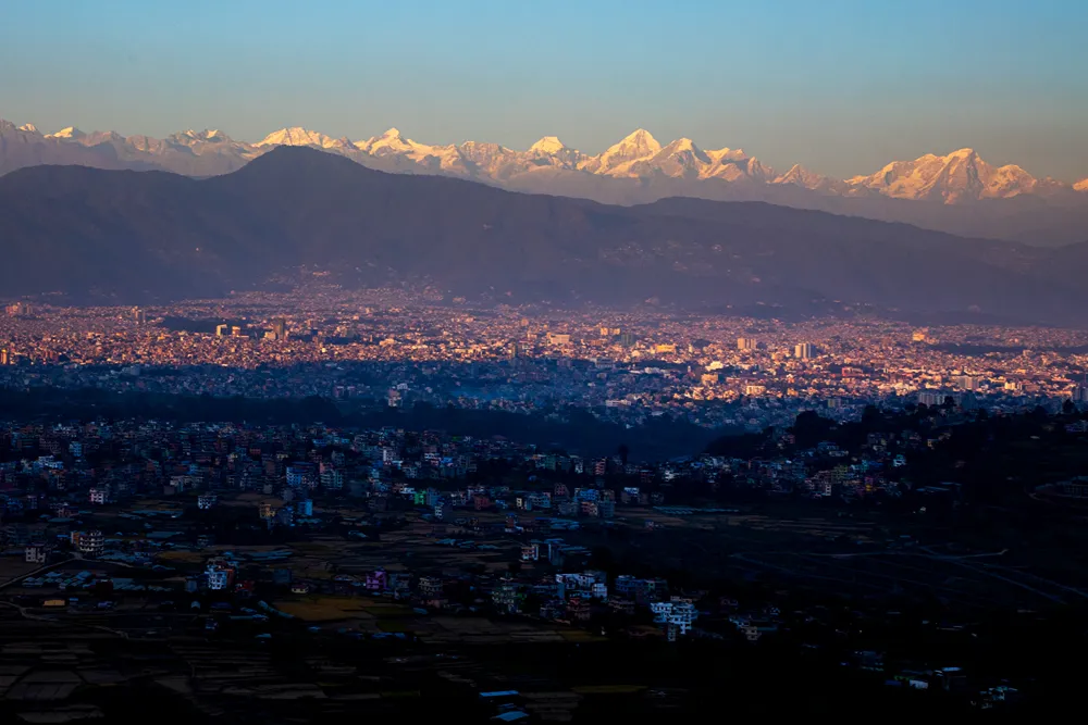Gün batımında, karlı dağların eteklerinde uzanan yoğun yerleşimli bir şehir manzarası